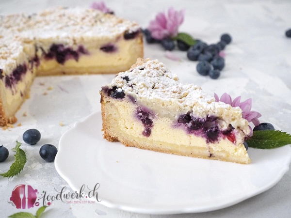 Ein Stück Blaubeer-Käsekuchen auf weissem Teller mit Blaubeeren.
